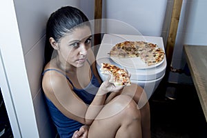 Bulimic woman feeling sick guilty sitting at the floor of the toilet leaning on WC eating pizza photo