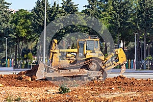 Bulldozer with a plow and a bucket in front levels the ground next to a city park