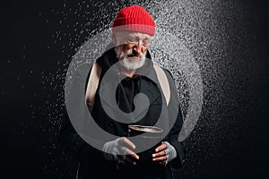 Bum with grey beard stand under heavy rain with cup for money