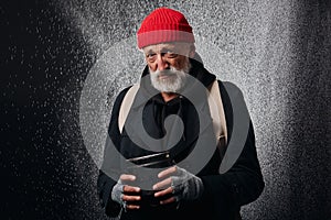 Bum with grey beard stand under heavy rain with cup for money