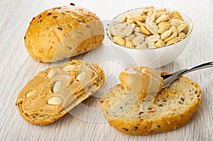 Bun, peanut in bowl, sandwich with peanut butter, spoon with paste on bread on table