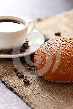 Bun with sesame seed, white mug filled coffee on saucer and scattered coffee beans on burlap tablecloth top view