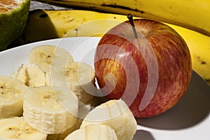 A bunch of bananas, a sliced banana, and an apple on a plate on a table. Selective focus