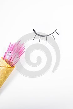 Bunch of pink eyelash extension brushes folded into ice cream cone on white background with pattern of closed eye.