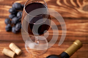 A bunch of ripe blue grapes with a glass of red wine with wine corks nearby, and a sealed bottle of red wine on a textured wooden