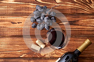 A bunch of ripe blue grapes with a glass of red wine with wine corks nearby, and a sealed bottle of red wine on a textured wooden