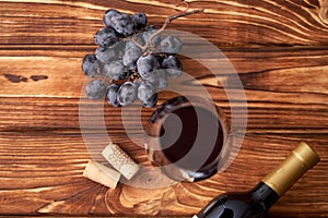 A bunch of ripe blue grapes with a glass of red wine with wine corks nearby, and a sealed bottle of red wine on a textured wooden