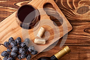 A bunch of ripe blue grapes with a glass of red wine with wine corks nearby, and a sealed bottle of red wine on a textured wooden