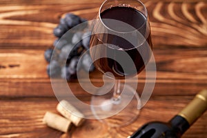 A bunch of ripe blue grapes with a glass of red wine with wine corks nearby, and a sealed bottle of red wine on a textured wooden
