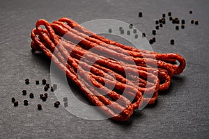 Bunch of thin dry smoked sausage kabanos or cabanossi and pepper grains on a black stone serving board. Traditional polish meat