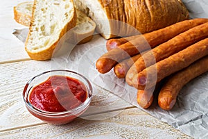 Bunch of thin smoked sausages, ketchup and slices of bread on a white wooden table. Sausage and fresh vegetables as a source of