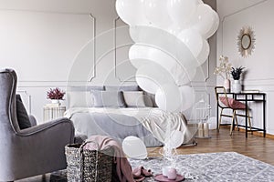 Bunch of white balloons in the middle of classy grey bedroom with elegant dresser with golden mirror, copy space on the empty wall