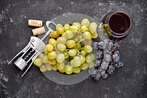 Bunches of ripe green and blue grapes with a glass of red wine, a corkscrew and wine corks on a dark stone background. Top view.