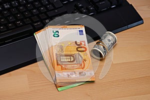 A bundle of Euro banknotes on a computer keyboard. Near a roll of one hundred dollars