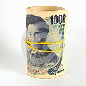 A bundle of Japanese 1000 one thousand yen bills stands on a white paper surface. Banknotes are rolled up and tied with an elastic