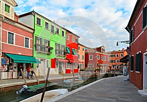Burano island picturesque street with small colored houses in row, water canal with fishermans boats, cloudy blue sky