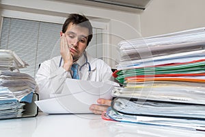 Bureaucracy in medicine concept. Tired overworked doctor is reading medical report. Many documents on desk