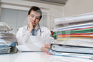 Bureaucracy in medicine concept. Tired overworked doctor is reading medical report. Many documents on desk photo