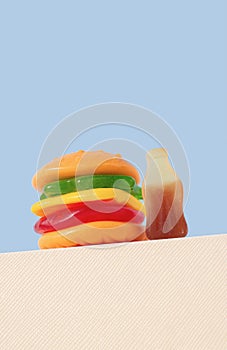 Burger and bottle shape gummy candy on white podium, blue background. Sweet food concept