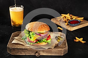 Burger with French fries and beer. Hamburger with beef, cheese, onion, tomato, and green salad, on a dark background