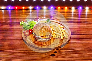 Burger with French fries on a round board on a wooden table with multicolored lights on the background