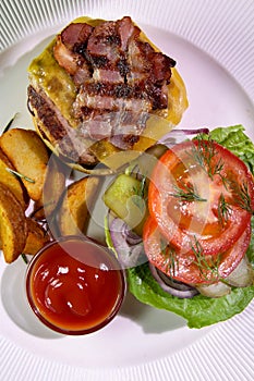 Burger with fried bacon, french fries, vegetables and ketchup on a white plate.View from above.Close-up.Caloric intake