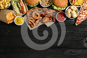 Burgers, onion rings and other fast food on black wooden table, flat lay with space for text