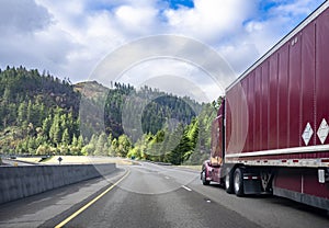 Burgundy big rig classic semi truck with dry van semi trailer running on the winding mountain highway road