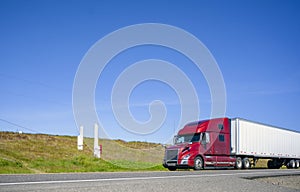 Burgundy bonnet big rig semi truck transporting cargo in dry van semi trailer driving on the road with hillside