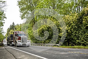 Burgundy classic big rig semi truck tractor with heavy grille guard transporting covered cargo on flat bed semi trailer running on