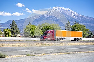 Burgundy long hauler big rig semi truck with orange dry van semi trailer driving on the flat highway road with snow mountain on