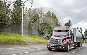 Burgundy low cab big rig semi truck transporting cargo in low profile covered semi trailer running on the multiline highway road