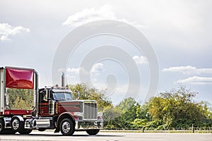 Burgundy old style day cab big rig semi truck tractor with high chrome pipes transporting cargo in bulk semi trailer with red