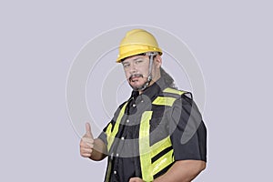A burly foreman or engineer wearing a hardhat and safety vest gives a thumbs up of approval. Isolated on a gray background