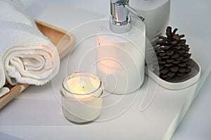 Burning aromatic candle, towels and bottles on white table.