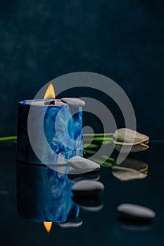 Burning blue handmade candle. against the background of a flower and stones, reflected in the glass.