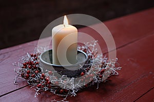 Burning candle in a bowl decorated with a small wreath from rose hips and silver cushion bush on a red wooden table, winter