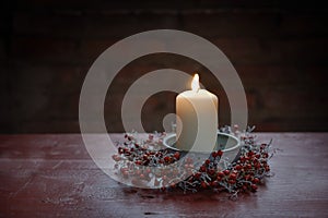 Burning candle in a bowl with a small wreath from rose hips and silver cushion bush on a red wooden table, winter decoration for