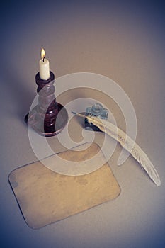 Burning candle in candlestick, letter paper blank, quill pen, inkwell on the table. Vintage style filtered photo