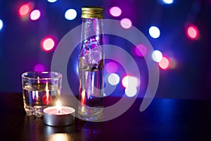 Burning candle with a drink on the background with colorful lights. Nearby is a glass and a bottle of drink. Holiday accessories