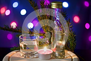 Burning candle with a drink on the background with colorful lights. Nearby is a wineglass, a bottle of drink and a fir branch.