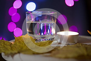 Burning candle and a glass with a drink on the background with colorful lights. Nearby are autumn maple leaves. Holiday