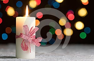 Burning candle with red bow, in snow, with defocussed fairy lights, bokeh in the background