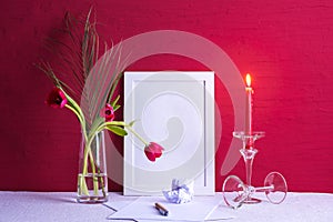 Burning candle, writing materials, fern leaf and tulips in a vase, white photo frames on a red background