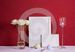 Burning candle, writing materials, white roses in a glass vase, white photo frames on a red background