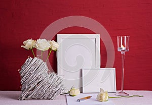 Burning candle, writing materials, white roses in a vase, white photo frames, wooden heart on a red background