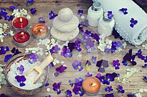 Burning candles and spa set with bath salt, stones for hot massage and skin lotions in the midst of small purple flowers of