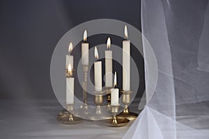 Burning candles in vintage metal candlesticks on white table against abstract white tulle material  background.
