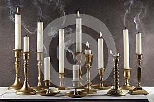 Burning and extinguished candles with smokes in vintage metal candlesticks on white wooden table against dark stone background.