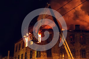 Burning house at night, roof of building in flames of fire and smoke, firefighter on crane extinguishes fire with water from hose